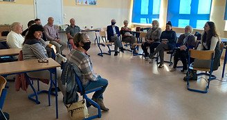 Zdjęcie zrobione w klasie w szkole. Przedstawia starsze osoby siedzące na krzesłach w półkole na środku sali. Najbardziej na prawo widać młodą kobietę, która prowadzi zajęcia. 